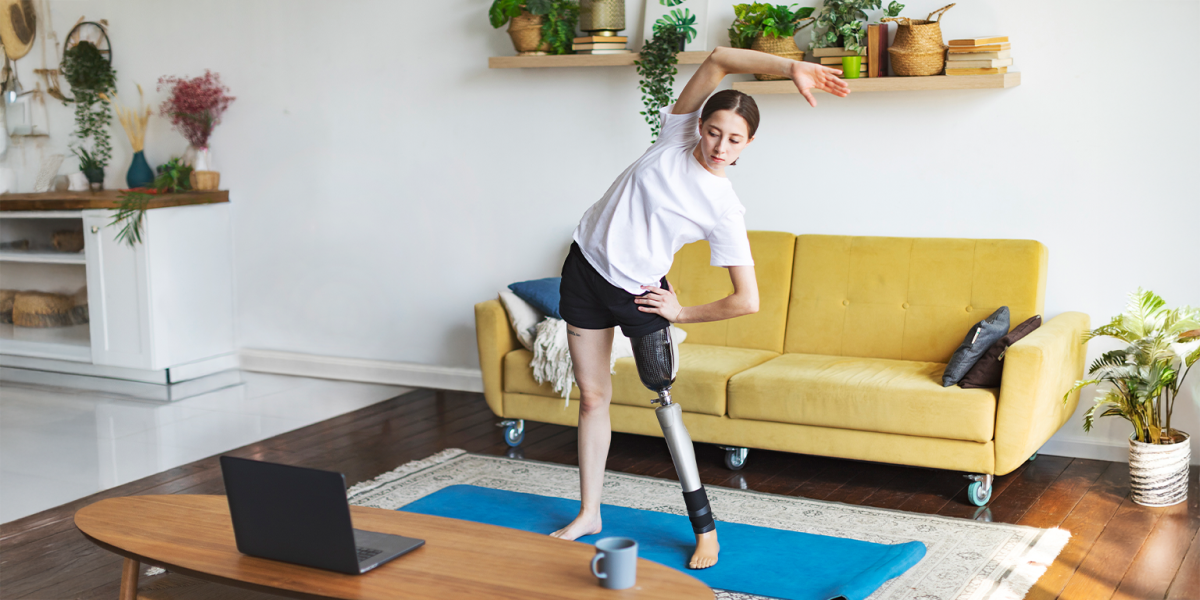 A woman with a prosthetic leg performs a side stretch on a blue yoga mat in a bright living room while following a workout on a laptop placed on a nearby coffee table.