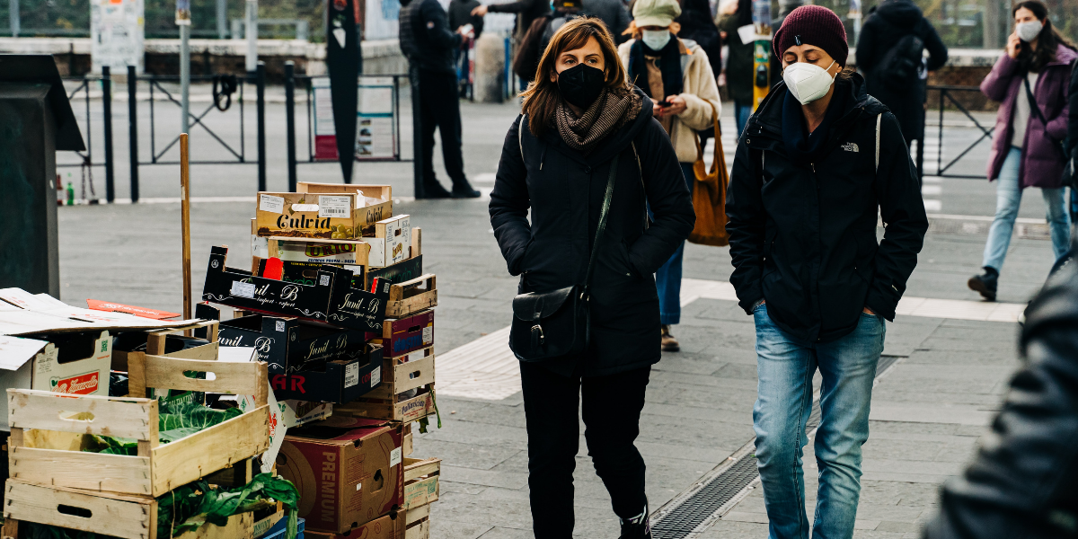 Several people walking around a large city with their face masks on. On the left side of the image, people are walking past stacked crates of vegetables.