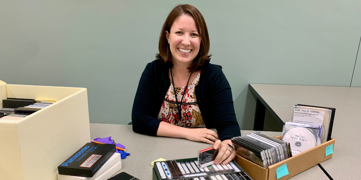 A woman smiling at the camera sitting at a table with cassettes and videotapes