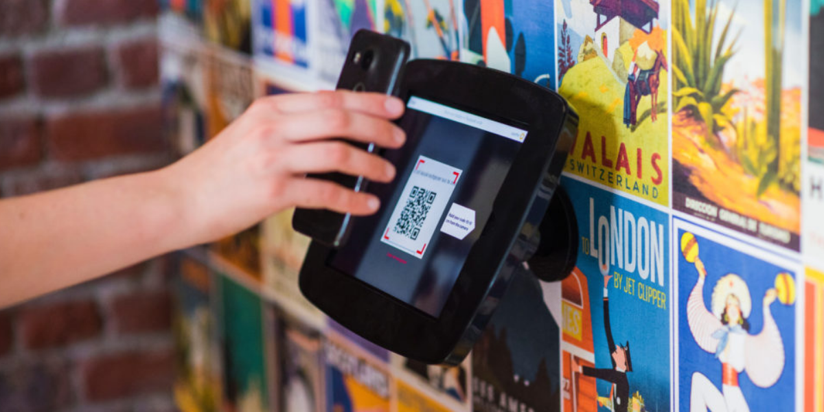 A person holding their smartphone up to a kiosk with a QR code on the screen. The kiosk is pinned on a wall with several colorful posters.