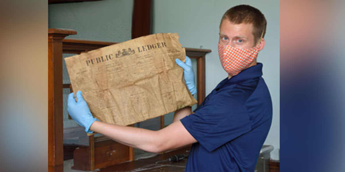 man with face mask on and gloves holding a discolored newspaper