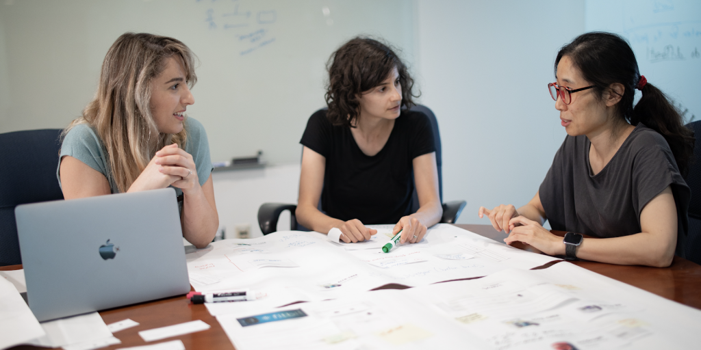 Three women sitting around a table with markers, paper and a laptop on it